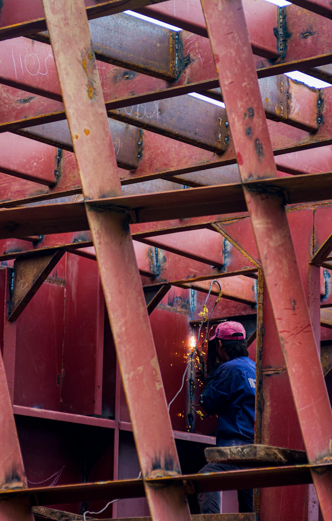A welder in action working on a large red metal framework, sparking visible.