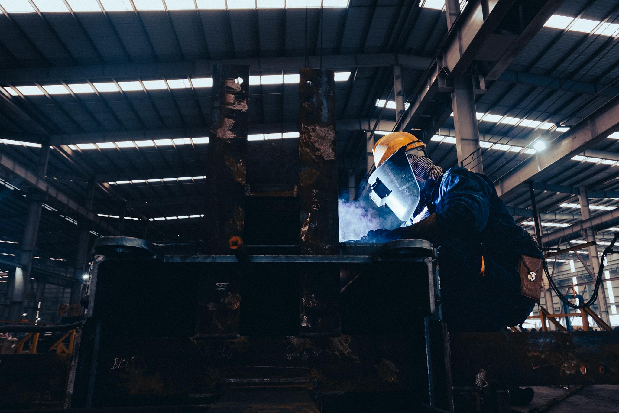 A welder wearing protective gear works on metal in a dimly lit industrial workshop.